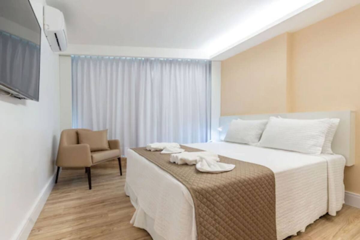 Ein Hotelzimmer mit Bett, Sessel, Fernseher und einem fenster mit Vorhängen.