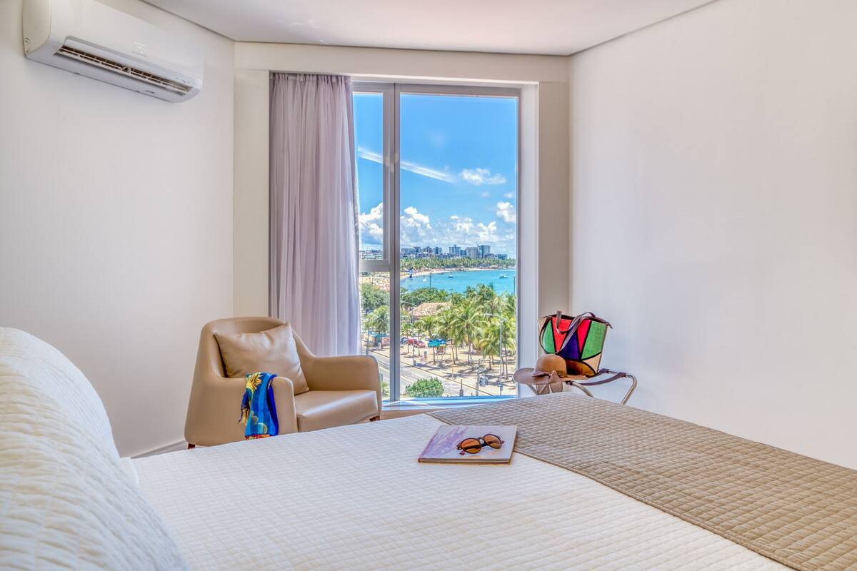Ein Hotelzimmer mit großem Fenster, einem Bett, einem Sessel und einem kleinen Tisch mit Buch und Sonnenbrille.