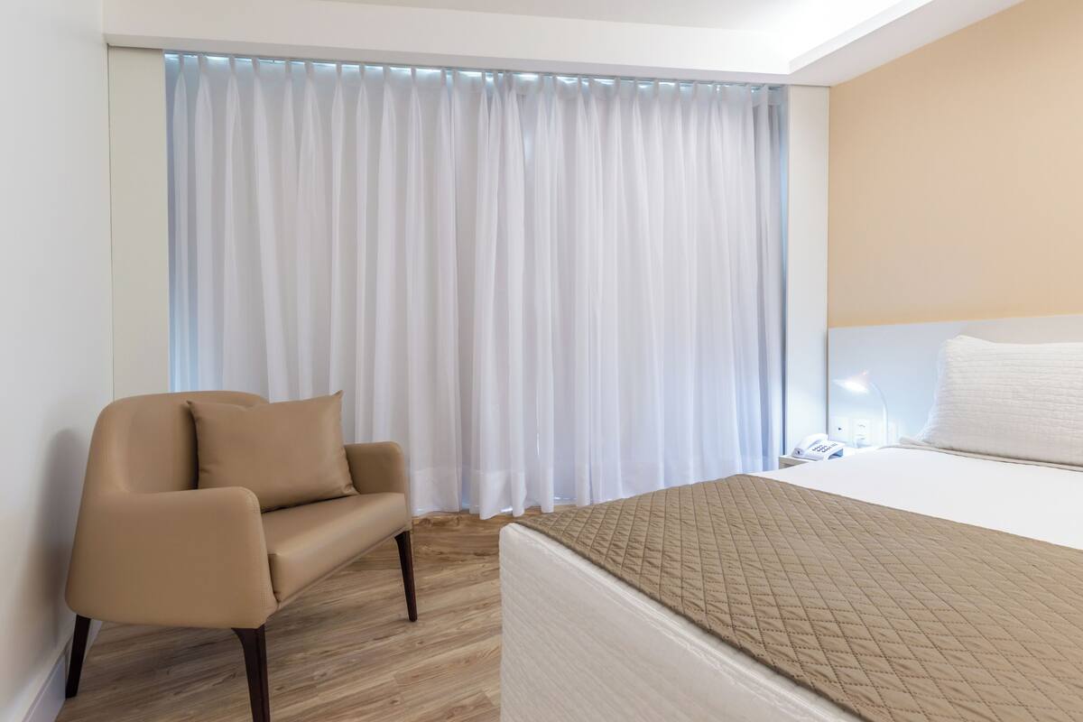 Ein Hotelzimmer mit Bett, Stuhl, Nachttisch und einem Fenster mit Vorhängen.