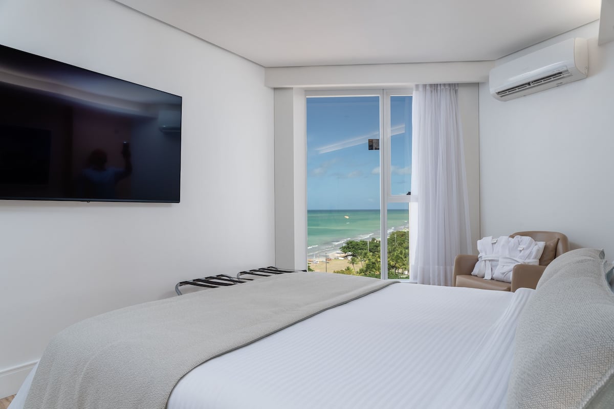 Ein Hotelzimmer mit Bett, einem an der Wand befestigten Fernseher, einem Sessel mit Bademantel und Blick auf das Meer durch ein großes Fenster.