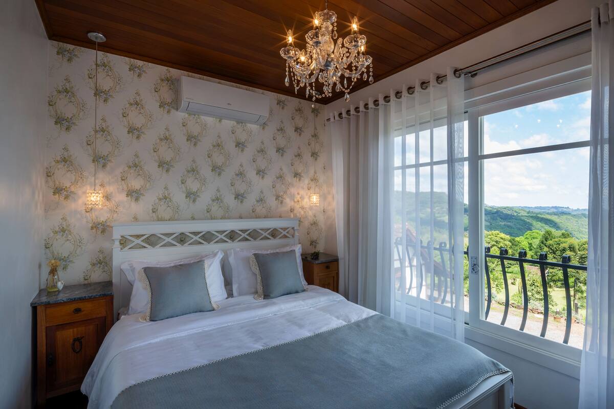 A bedroom with a large bed, a chandelier, a view of greenery through the window, and a wooden ceiling.