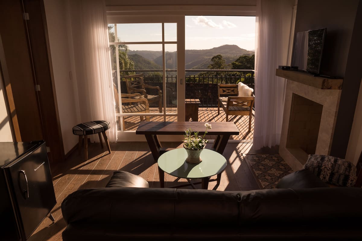 A living room with a sofa, a small table with a plant, a fireplace, and a balcony with a view of the mountains.