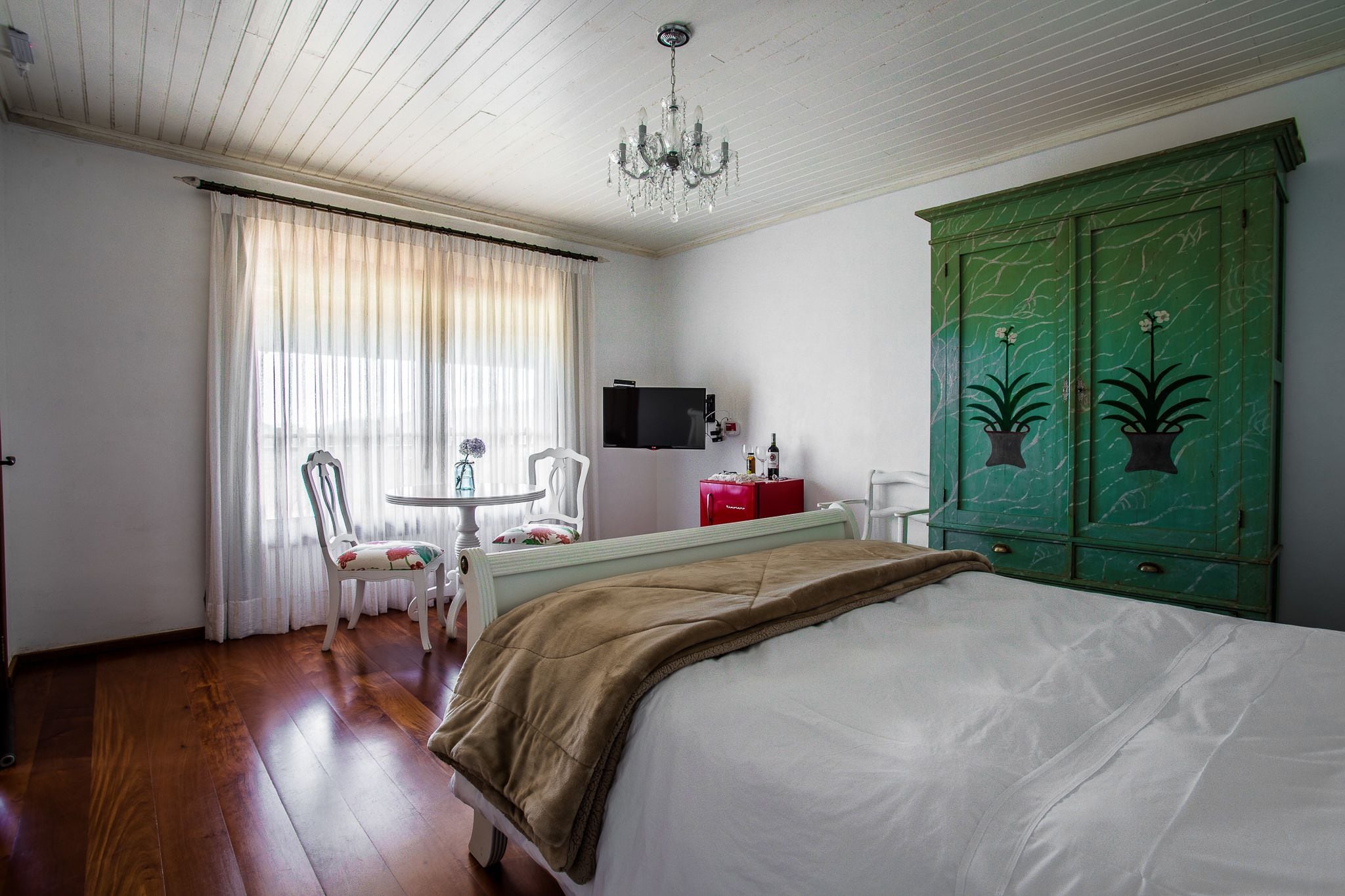 A bedroom with a bed, a green wardrobe, a television, a table with chairs, and a chandelier.