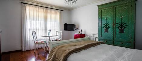 A bedroom with a bed, a green wardrobe, a television, a table with chairs, and a chandelier.