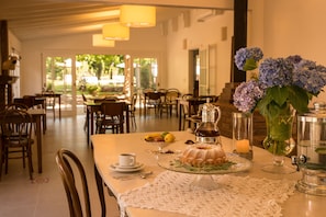 A dining area with a table set for a meal, including a cake and fruit. There are chairs, a vase with flowers, and a candle on the table.