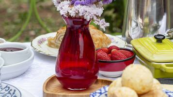 Uma mesa posta com um vaso vermelho, um coador de café de prata e diversos alimentos.