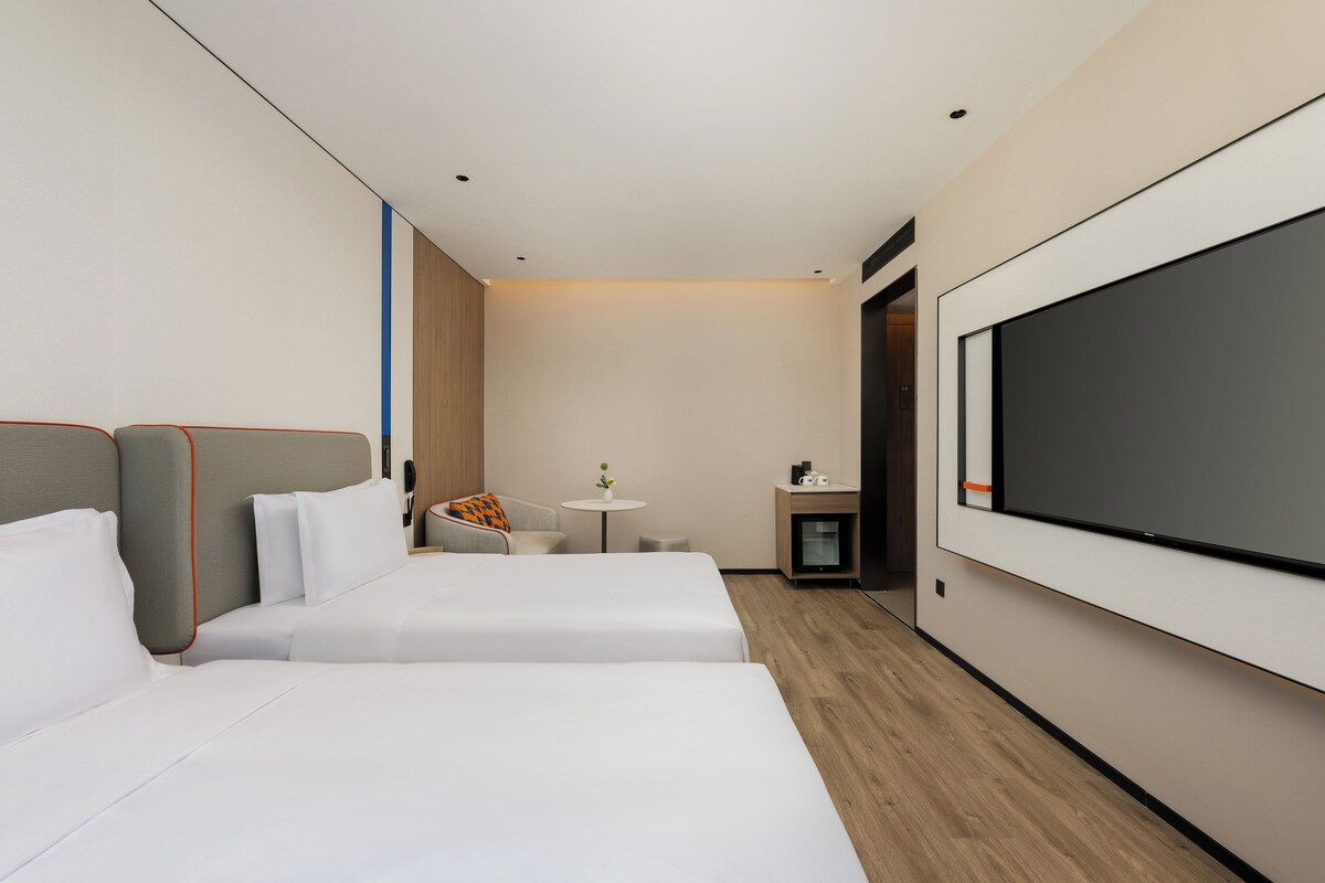 A hotel room with two beds, a small table with a vase, a TV, and a minibar.