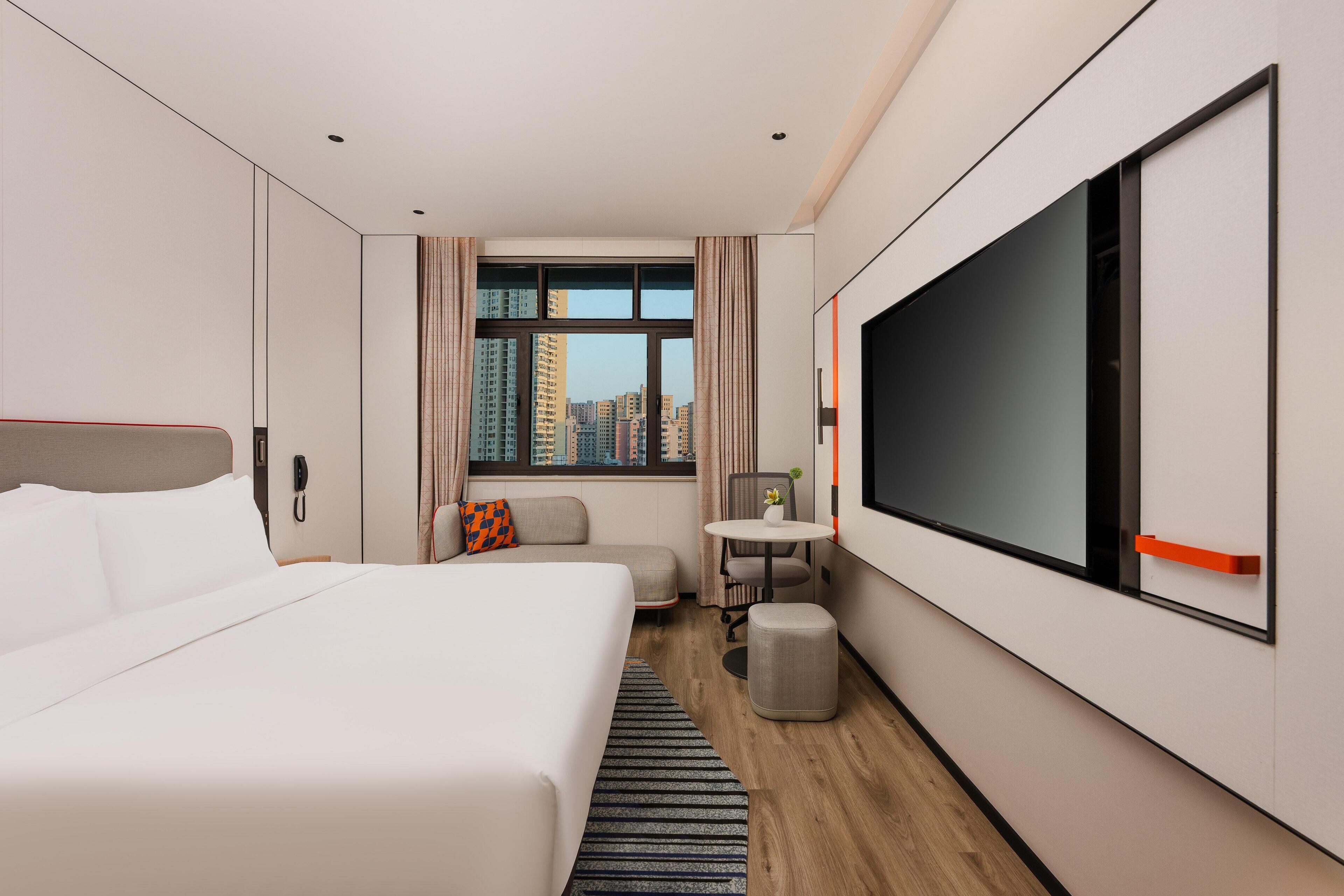 Habitación de hotel con una cama grande, un escritorio, una silla, una mesita, un televisor y una ventana con vistas a la ciudad.