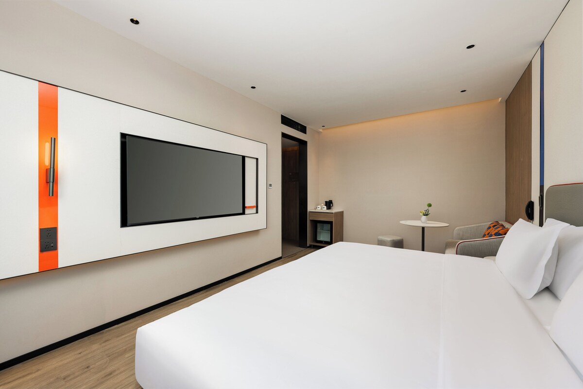 A modern hotel room with a large bed, a flat-screen TV mounted on the wall, a small round table with a plant, and a seating area with a sofa and armchairs.