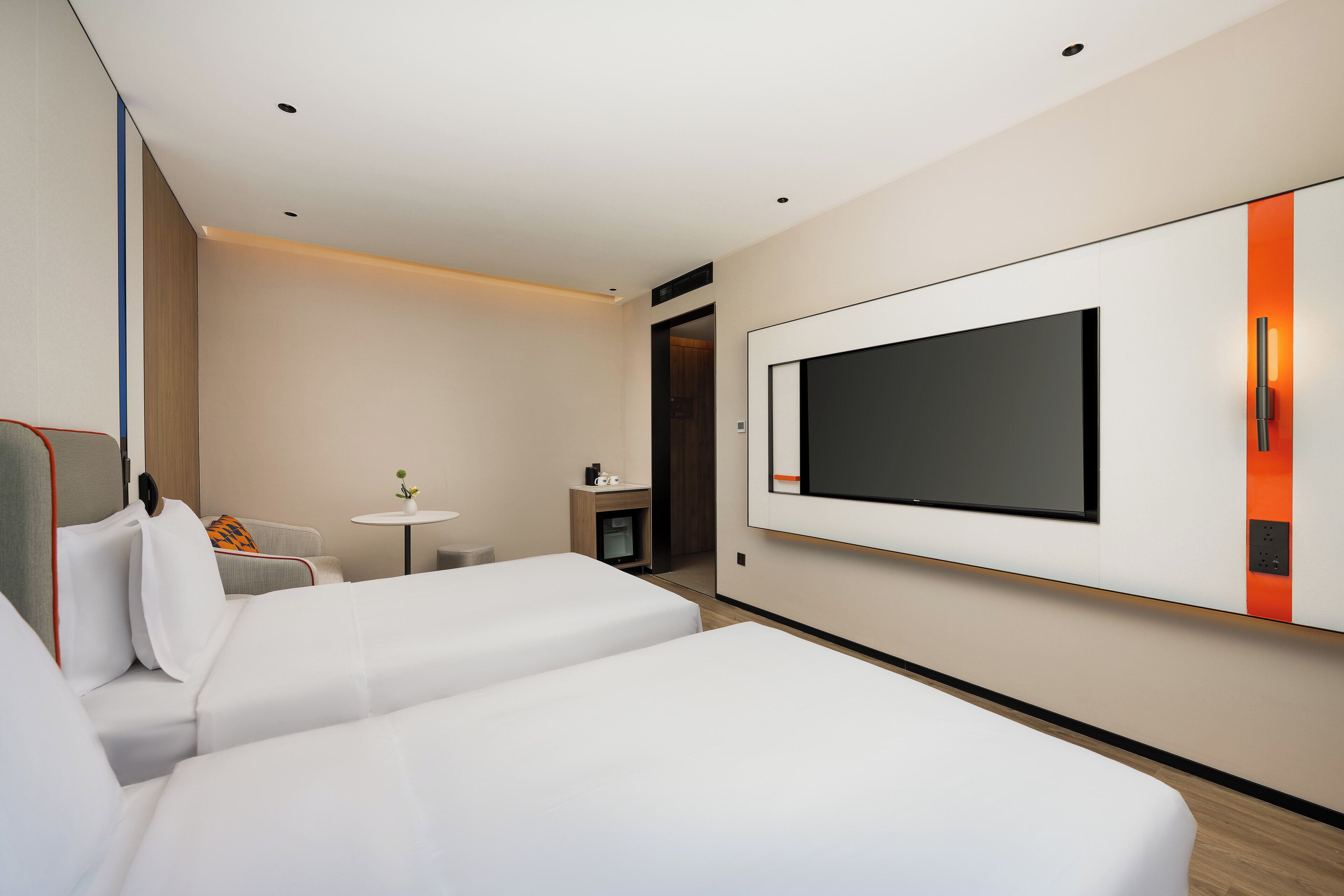 A hotel room with two beds, a TV mounted on the wall, a small table with a lamp, and a sofa with cushions.