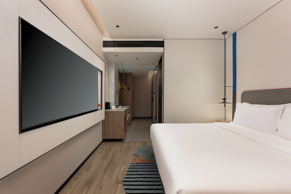 A modern hotel room with a large bed, a flat-screen TV mounted on the wall, a small desk, and a walk-in closet.