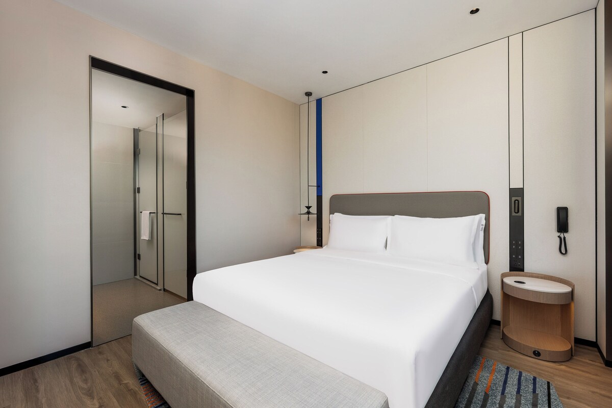 A modern hotel room with a large bed, a bedside table, a built-in bench, a mirror, and a phone on the wall.