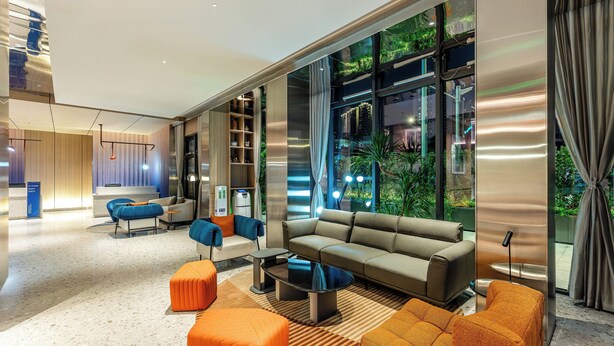 Una sala de estar moderna con un sofá, otomanas y una mesa de centro. Hay ventanas grandes con cortinas y vistas a la vegetación exterior.