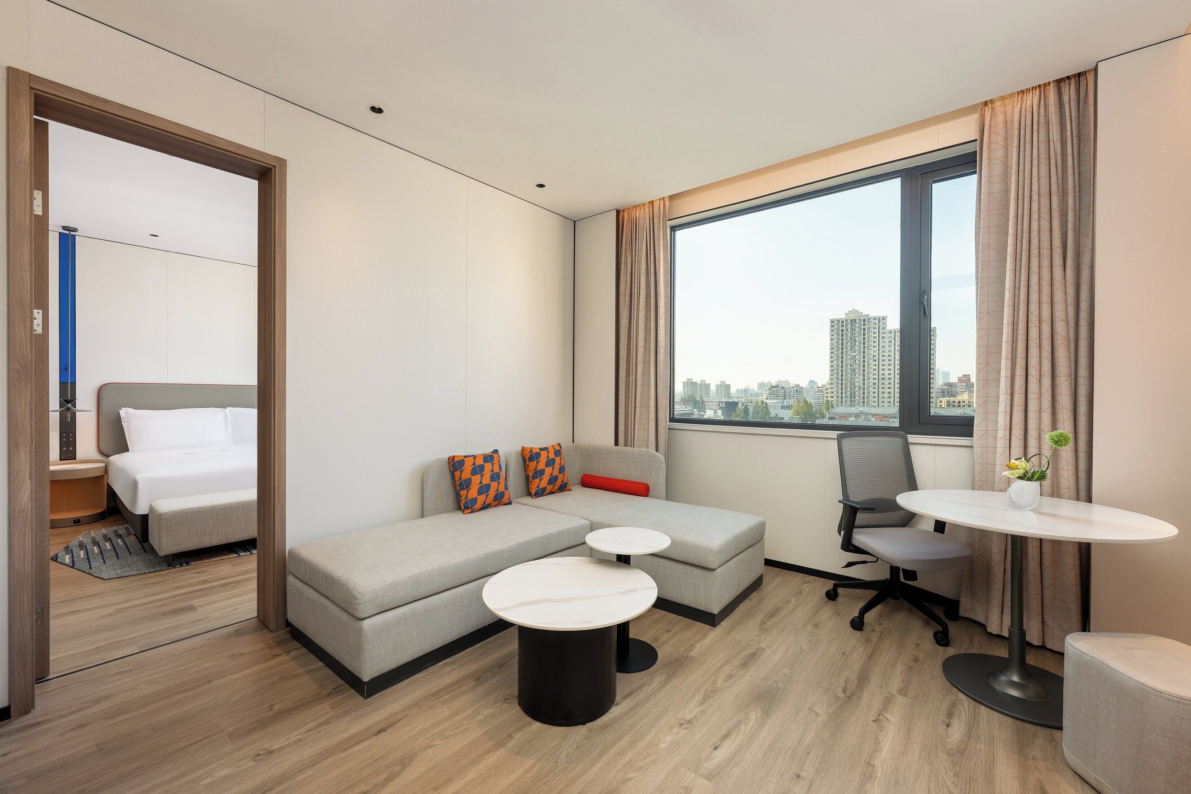 A modern hotel room with a bed, sofa, two round tables, and a large window with a city view.