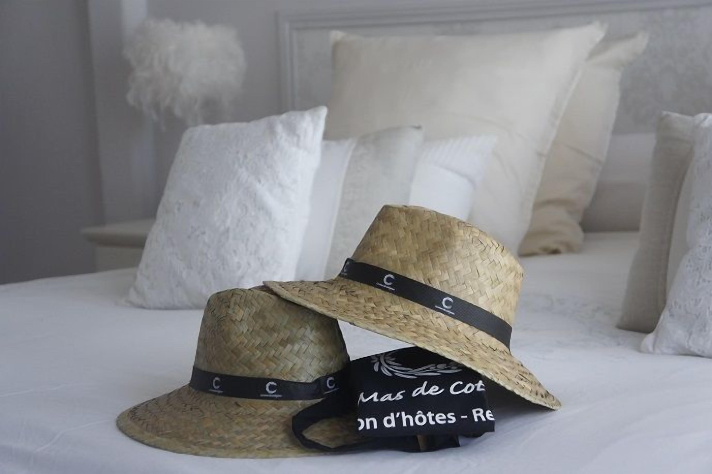 Deux chapeaux de paille ornés de bandeaux noirs, posés sur un lit recouvert de draps et d’oreillers blancs.