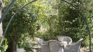 Een terras met rieten stoelen en een tafel, omgeven door groen en een metalen boog.
