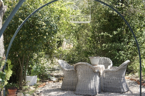 Un espace extérieur aménagé avec des chaises en osier et une table, entouré de verdure et d’une arche en métal.