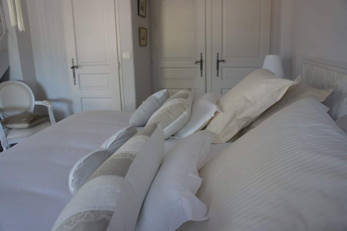 Un lit bien fait, avec du linge de lit blanc et plusieurs oreillers, une chaise blanche et une porte fermée, dans une pièce aux murs de couleur claire.