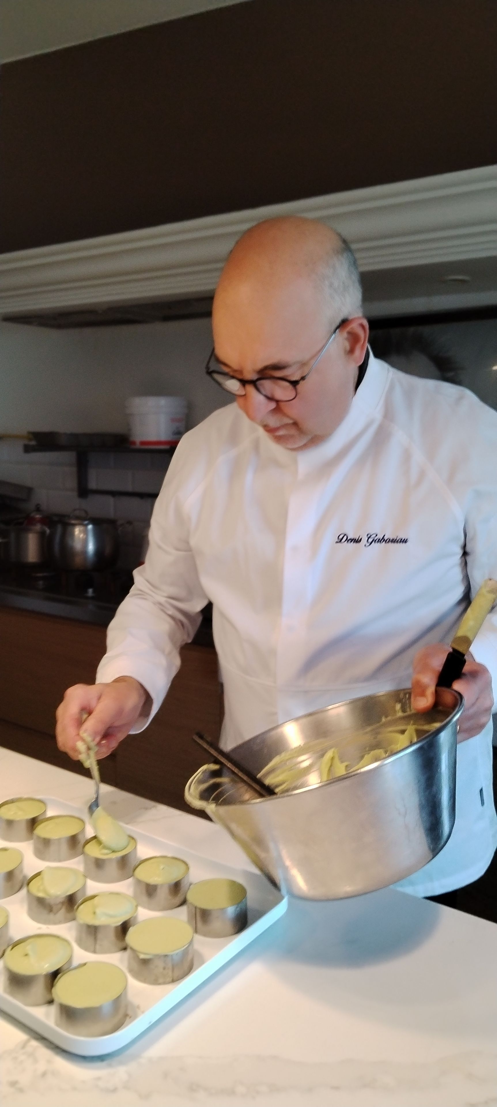Un chef con uniforme blanco sirve una mezcla amarilla en pequeños recipientes metálicos sobre una bandeja.