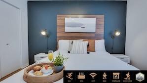Moderni hotellihuone, jossa on sänky, yöpöydät, yövalaisin, aamiaistarjotin ja seinälle ripustettu taulu.