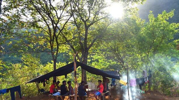 Eine Gruppe von Menschen sitzt in einem schattigen Bereich in bewaldeter Landschaft, im Hintergrund brennt ein Feuer.