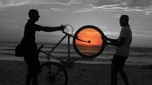 Zwei Personen halten ein Fahrrad und ein Rad vor einem Sonnenuntergang am Strand.