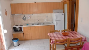 A compact kitchen with wooden cabinets, a sink, a stove, and a refrigerator. There is a dining table with chairs and a checkered tablecloth. A trash bin is visible near the kitchen entrance. - Anestis Apartments (Kavala)