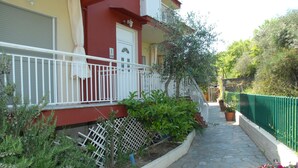 A narrow stone pathway leading to a red and white building with a white balcony, surrounded by greenery and potted plants. - Anestis Apartments (Kavala)