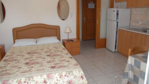 A bedroom with a bed, bedside table, lamp, and a view into the kitchen area. - Anestis Apartments (Kavala)