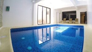 Ein Hallenbad mit klarem, blauem Wasser, Fliesenboden und Blick auf einen Balkon mit Sitzgelegenheiten.