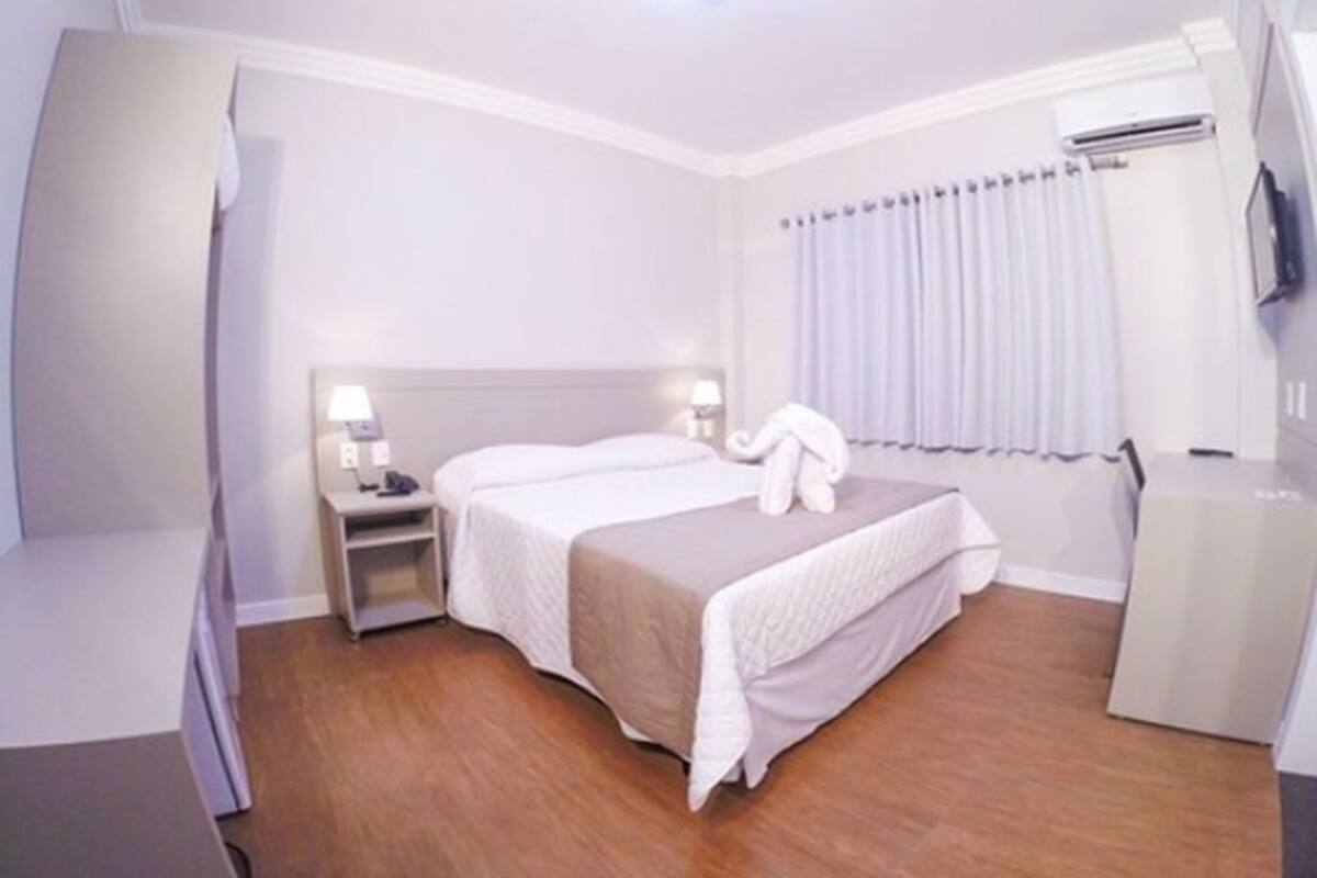 Una habitación de hotel con cama, mesitas de noche, lámpara, televisión y ventana con cortinas.