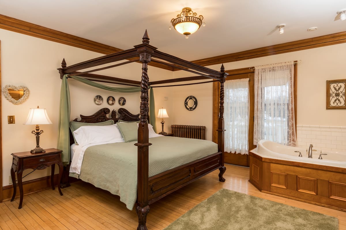 A four-poster bed with a green bedspread, a wooden floor, a green rug, a wooden nightstand with a lamp, a wooden dresser, and a large bathtub with a wooden base.
