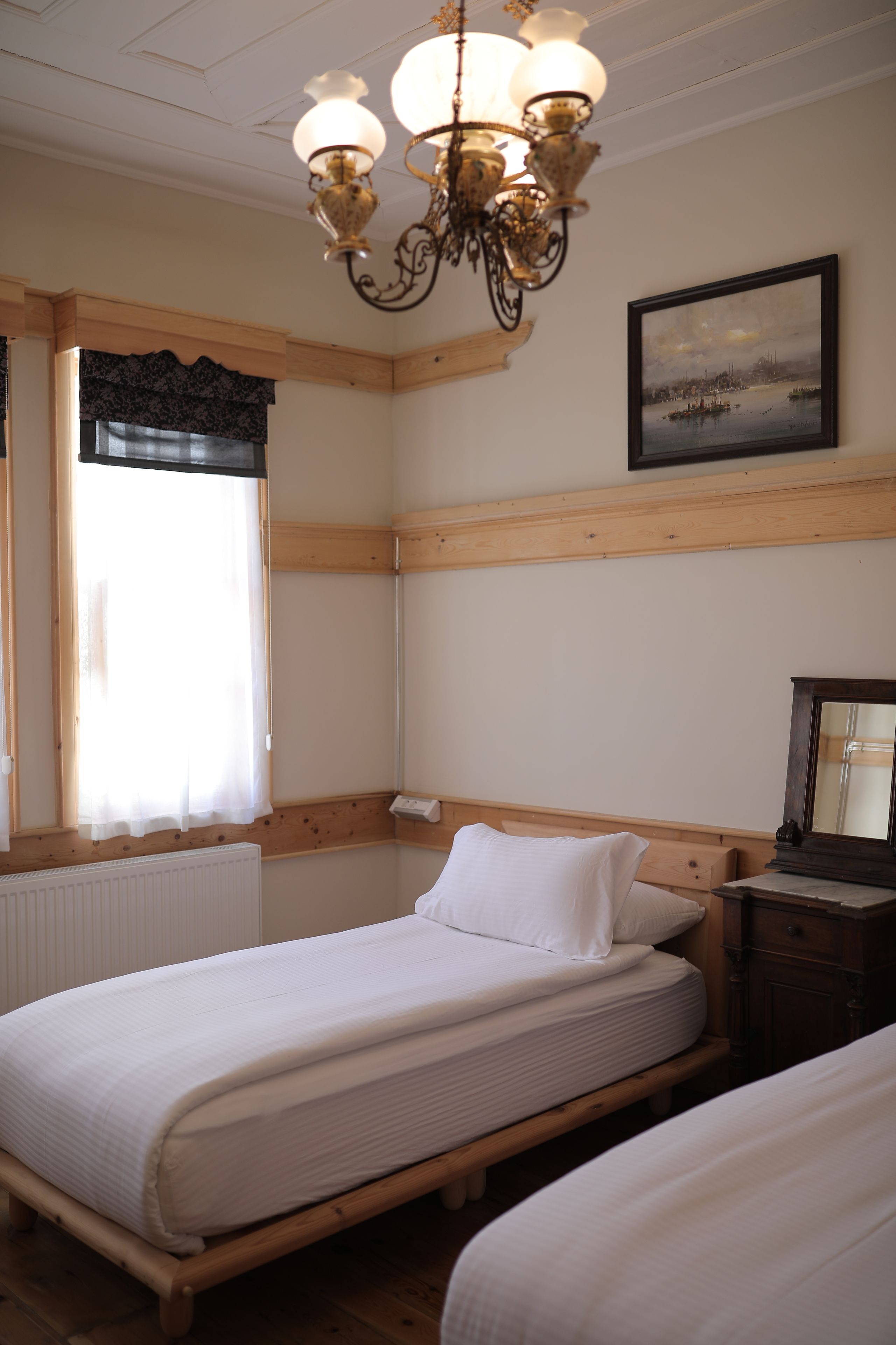 A bedroom with two single beds, a wooden headboard, a chandelier, a window with curtains, and a framed picture on the wall.
