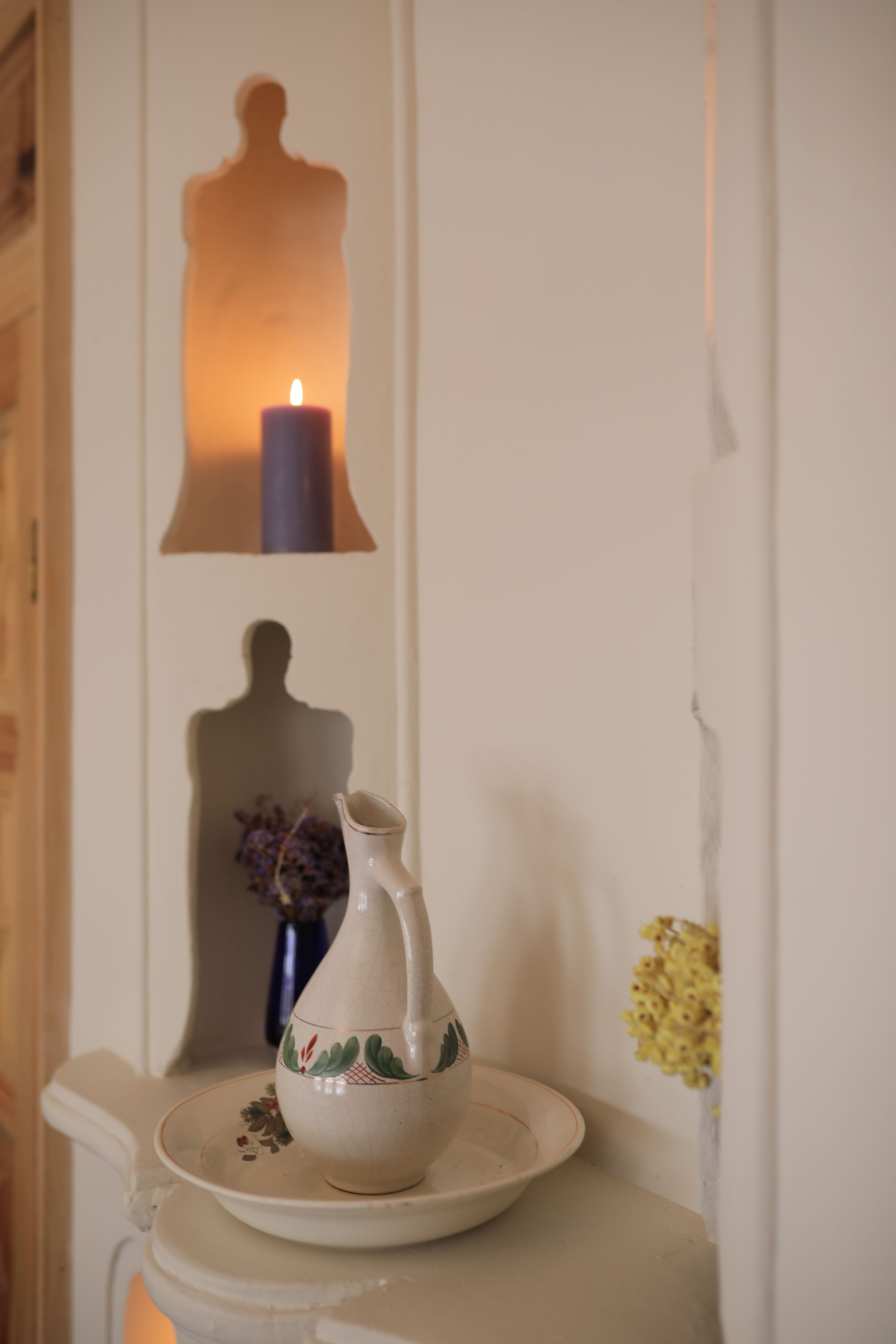 A white ceramic pitcher on a plate, a candle in a wall-mounted holder, and a shadow of a person on the wall.
