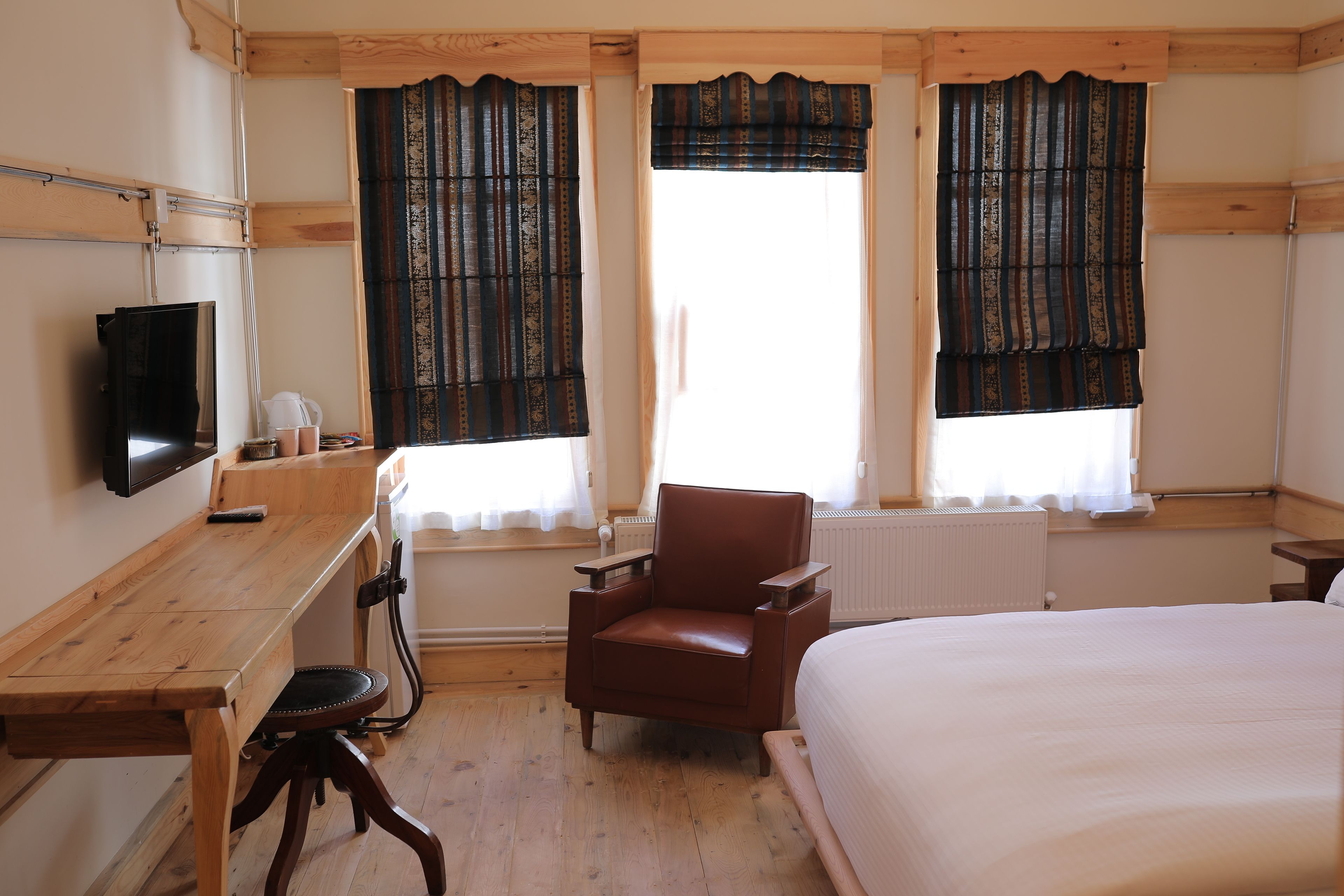 A room with a wooden desk, a brown leather chair, a bed, and two windows with patterned curtains.