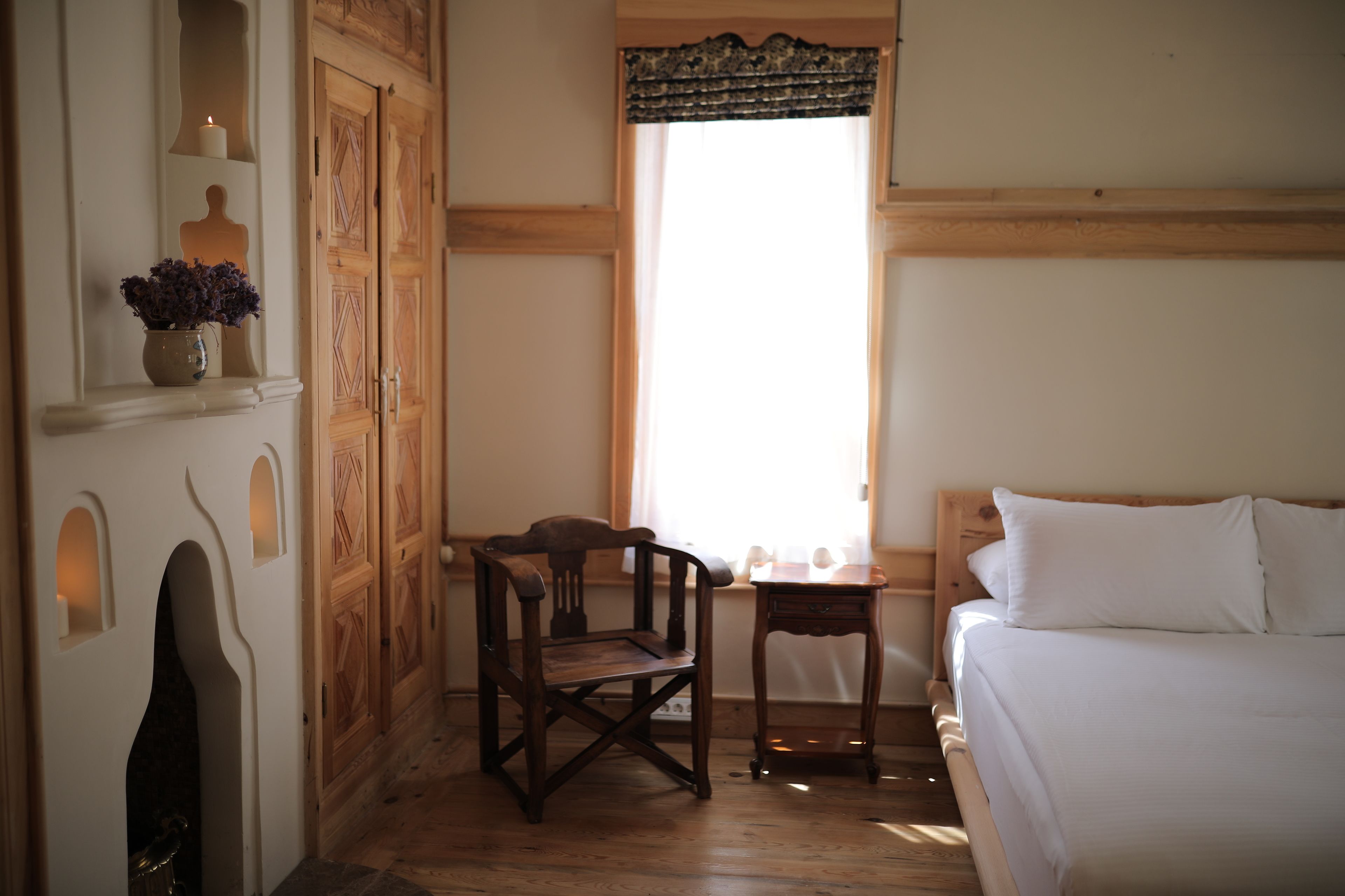 A room with a wooden bed, a small table, a chair, a window with curtains, and a wall-mounted shelf with a candle and flowers.