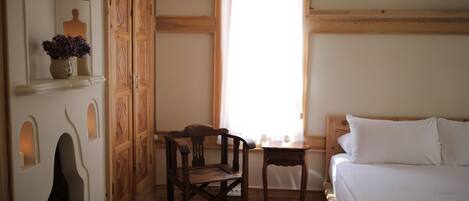 A room with a wooden bed, a small table, a chair, a window with curtains, and a wall-mounted shelf with a candle and flowers.