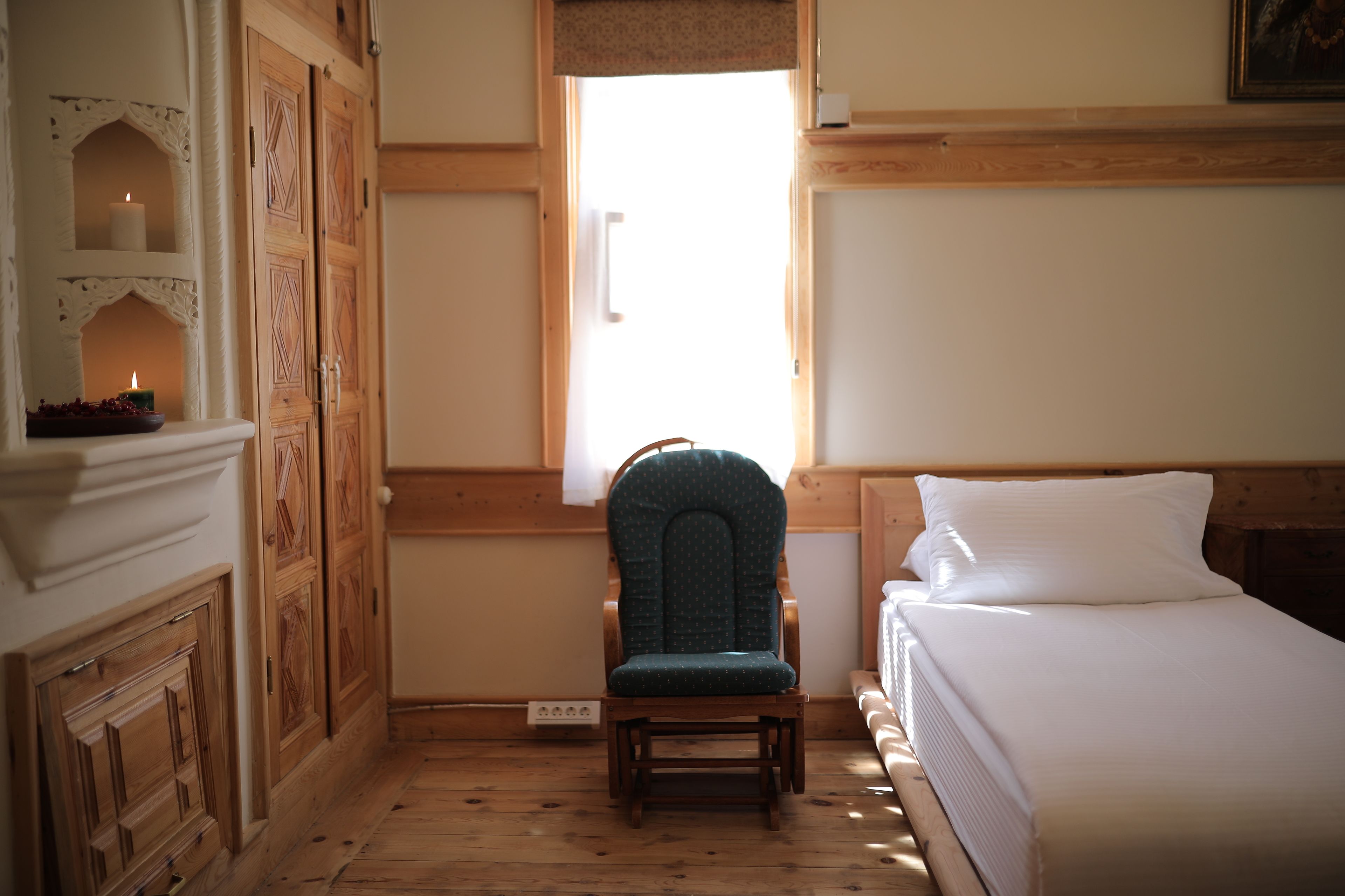 A bedroom with a wooden floor, a bed with white bedding, a green upholstered chair, and a window with a view of the outside.