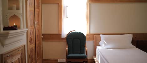 A bedroom with a wooden floor, a bed with white bedding, a green upholstered chair, and a window with a view of the outside.