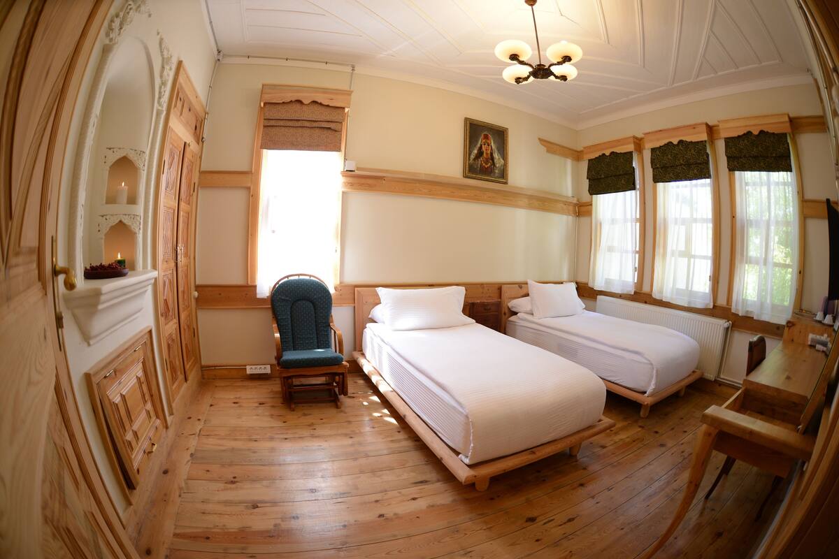A room with two beds, a chair, a wooden floor, a ceiling fan, and a window with curtains.