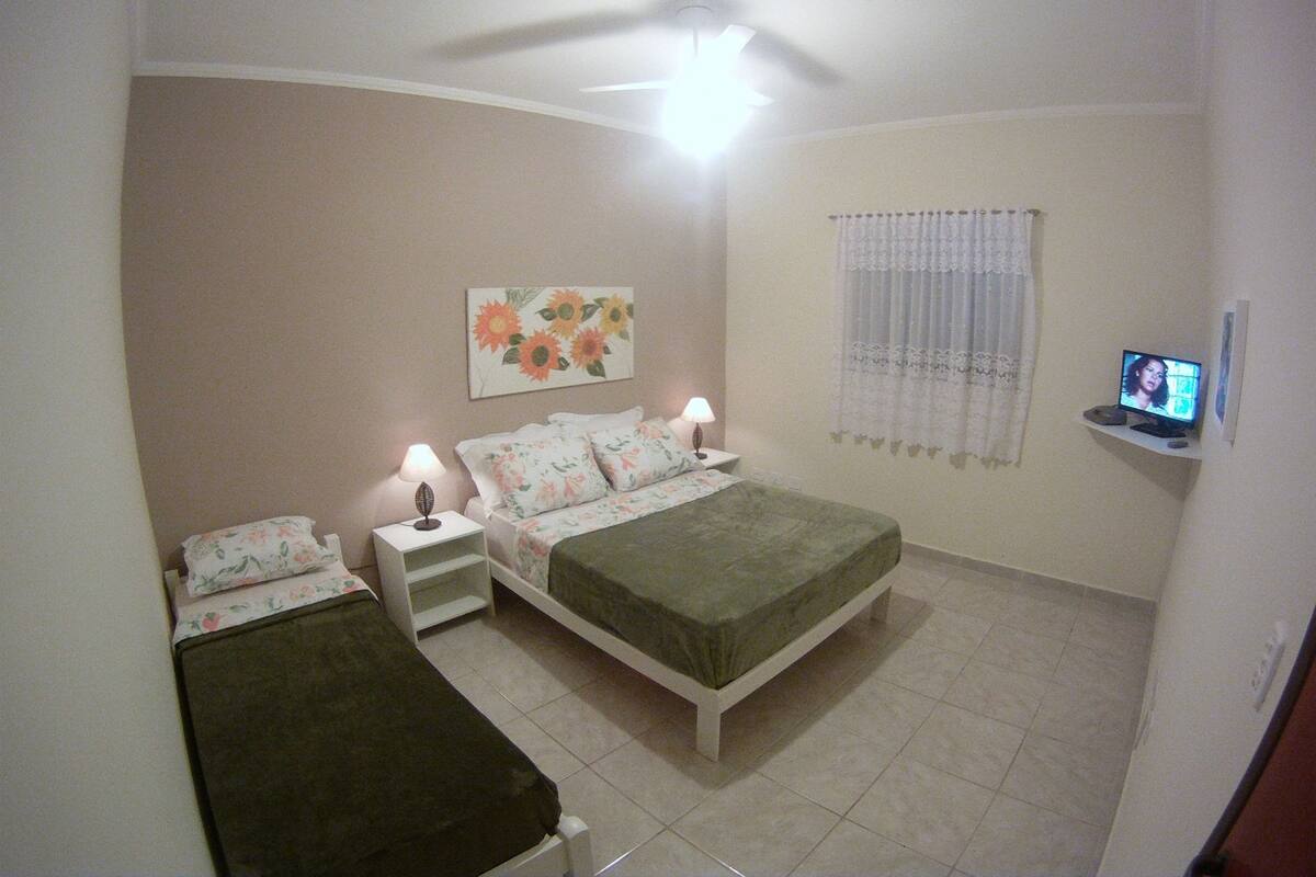 A bedroom with a bed, two bedside tables with lamps, a ceiling fan, a TV on a shelf, and a floral painting on the wall.