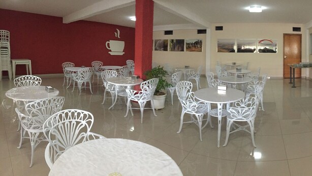 A hotel lobby with white metal chairs and tables, a buffet counter with various food items, and a red wall with a coffee cup symbol. - Pousada Golden House (Aguas de Sao Pedro)