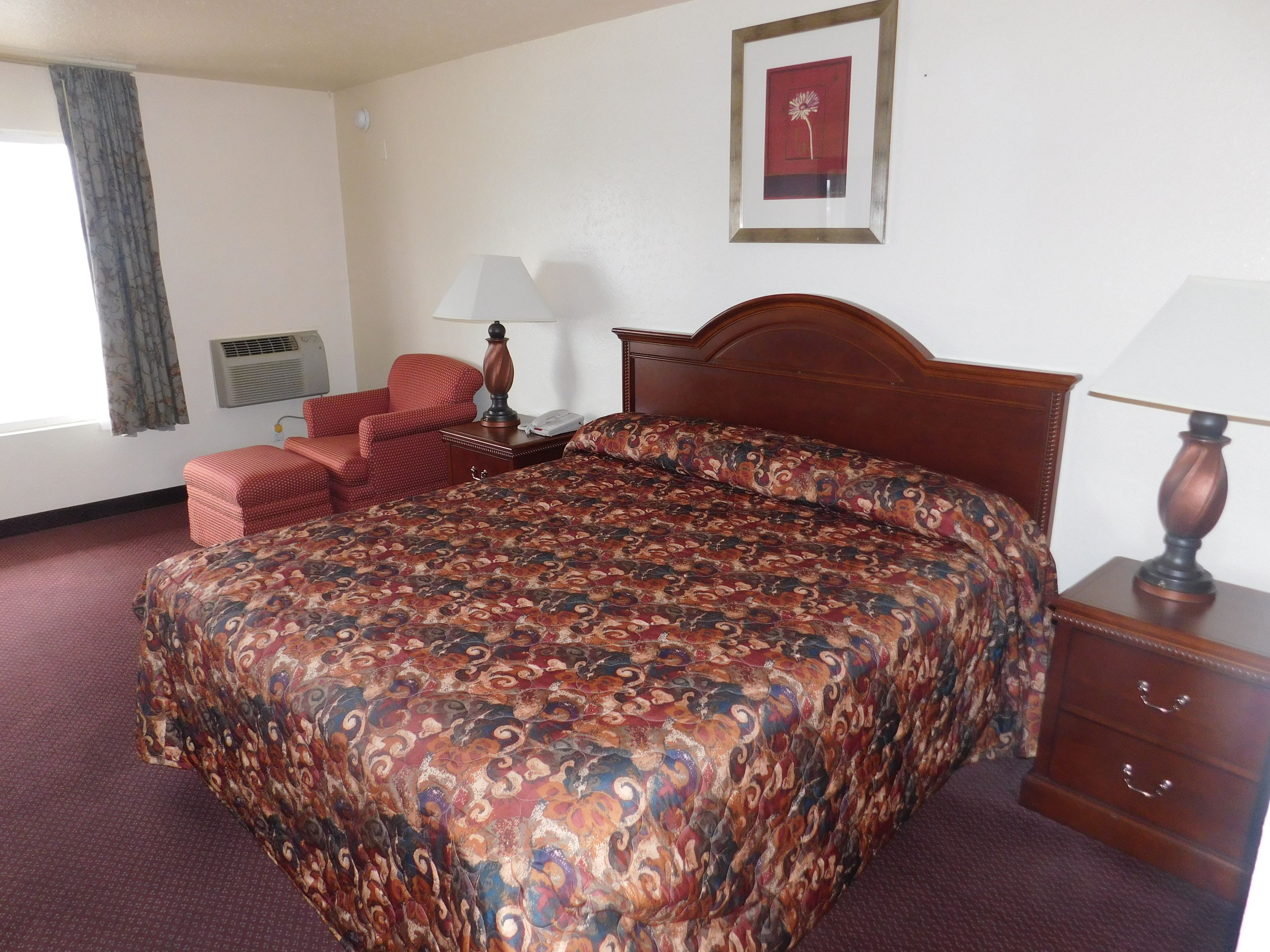 Een hotelkamer met een groot bed, een houten hoofdeinde, een rode fauteuil, een nachtkastje met een lamp en een ingelijste foto aan de muur.