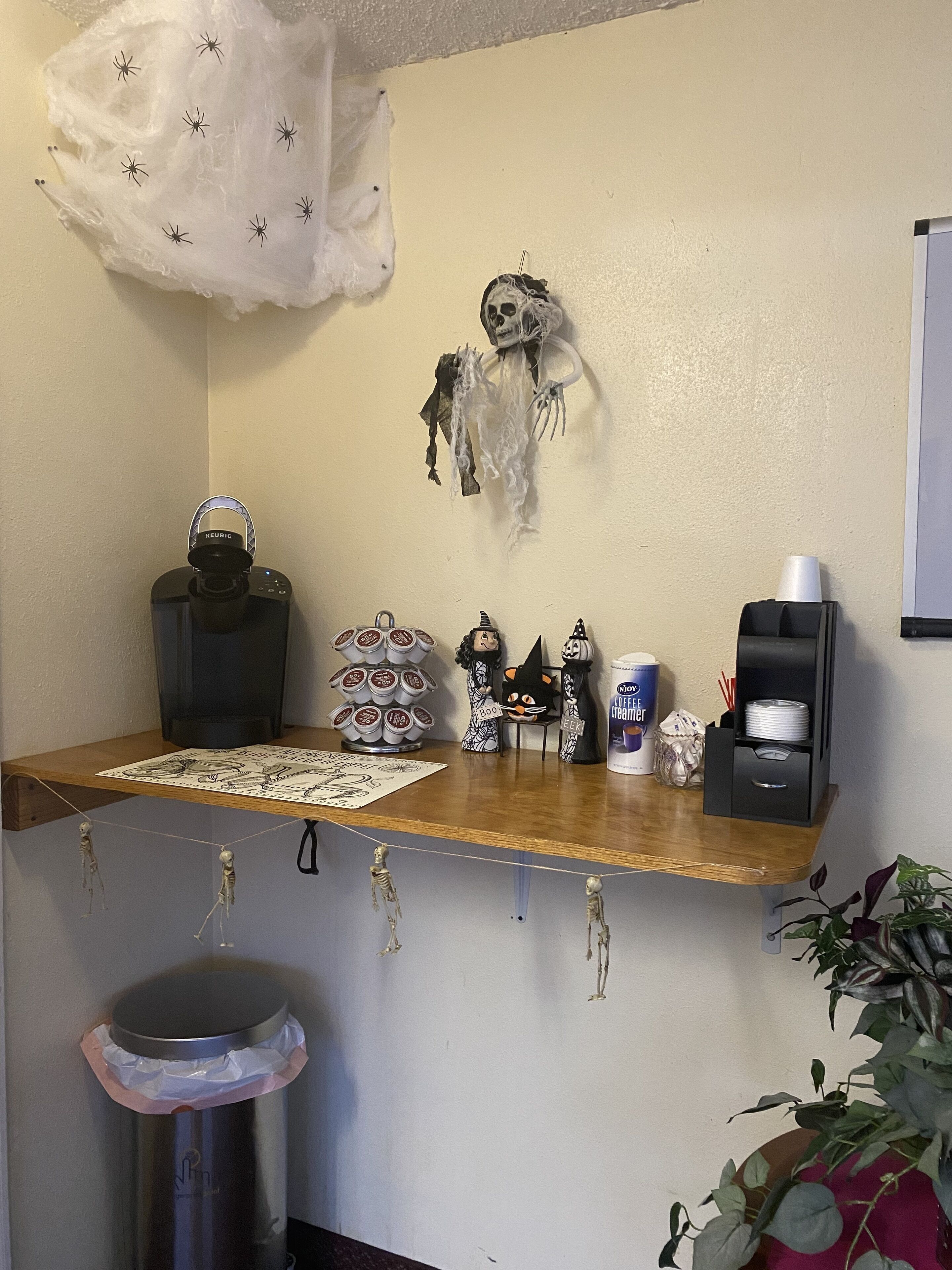 A coffee station with a Keurig machine, a coffee cup, a Monomail coffee creamer dispenser, and Halloween-themed decorations.