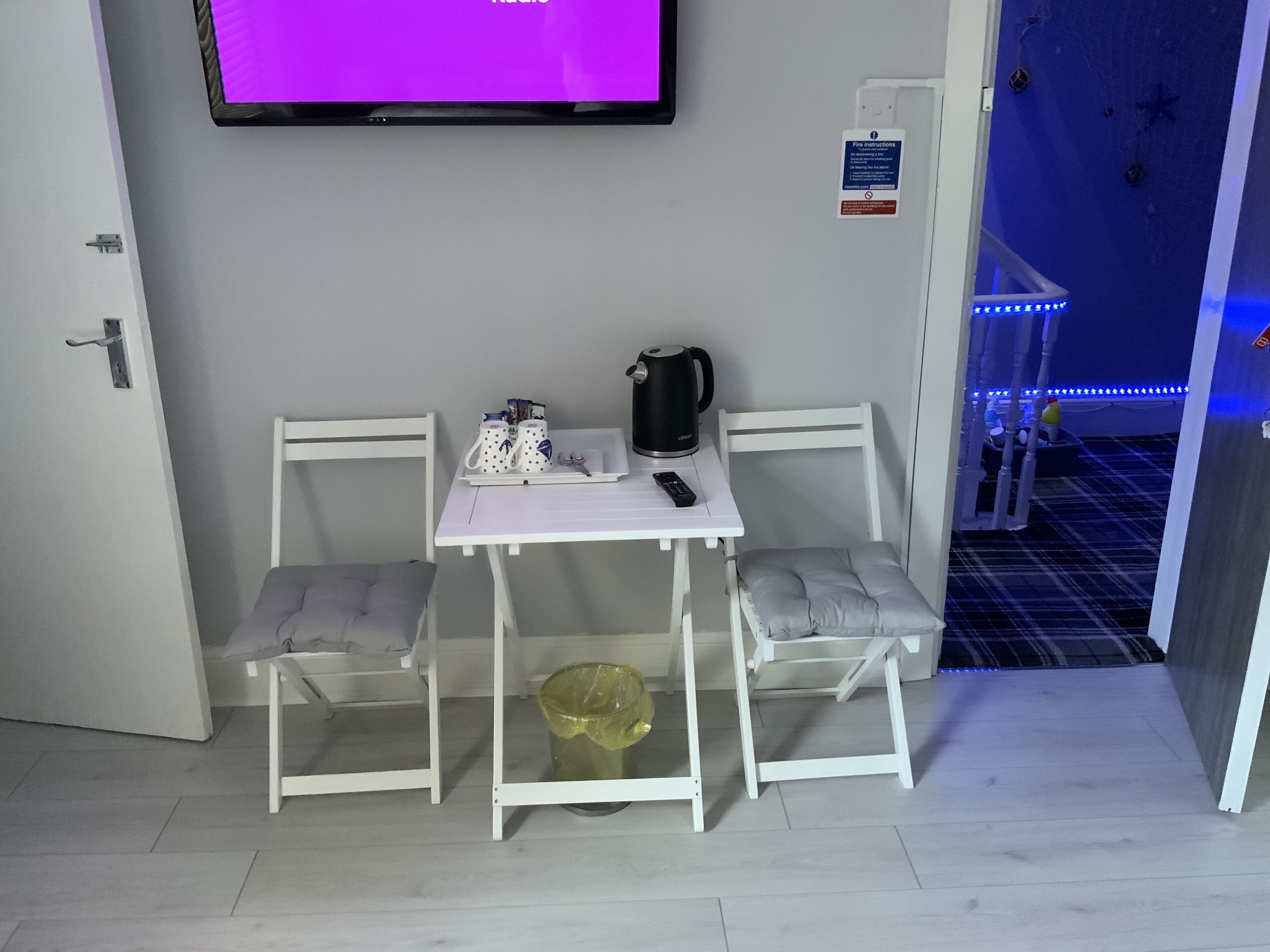Una stanza piccola con un tavolo bianco e due sedie, una TV fissata al muro e un cartello di sicurezza antincendio.