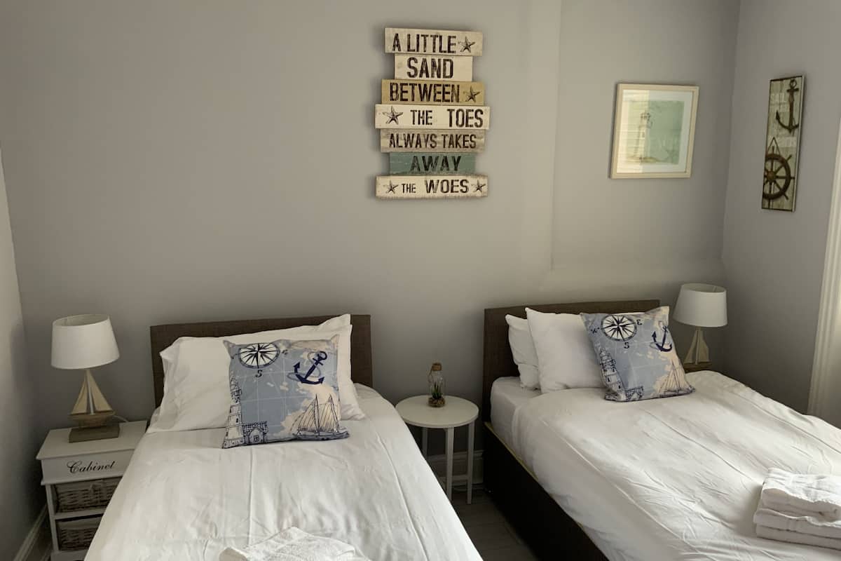 Quarto de hotel com duas camas, cada uma com roupa de cama branca e almofadas combinando. Há uma pequena mesa redonda entre as camas, e um armário com uma luminária no lado esquerdo.