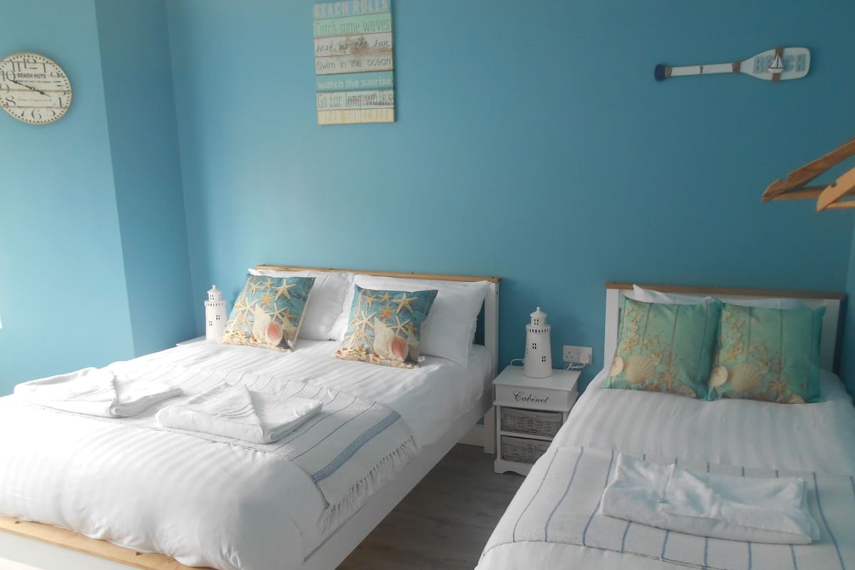 Quarto com duas camas, cada uma com cobreleira branca e almofadas a combinar. Há uma pequena mesa de cabeceira com luminária e um relógio de parede.
