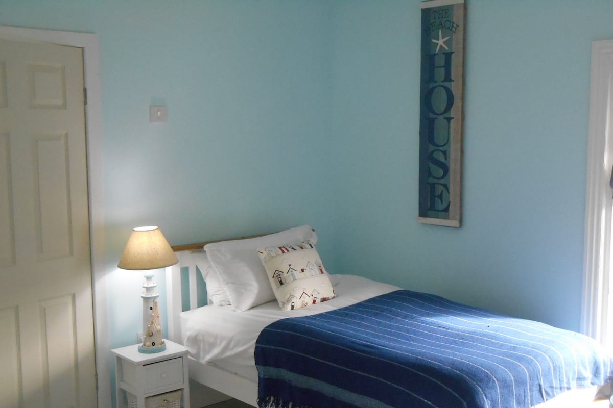 Quarto com uma cama, mesa de cabeceira com abajur, parede com letreiro que diz “HOUSE” e uma porta.