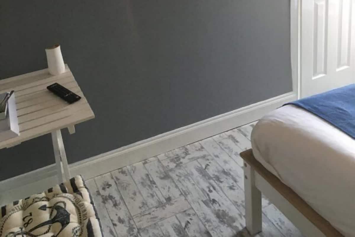 Um quarto com uma cama, uma mesinha com um controle remoto, uma prateleira com cabides e uma decoração de veleiro na parede.