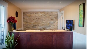 Un área de recepción con pared de piedra, mostrador de madera, un reloj, un jarrón con flores rojas y un cuadro en la pared.